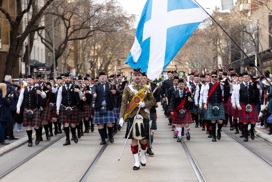 A parade of people wearing kilts, walking in formation down Collins Street, with the national flag of Scotland in the foreground.