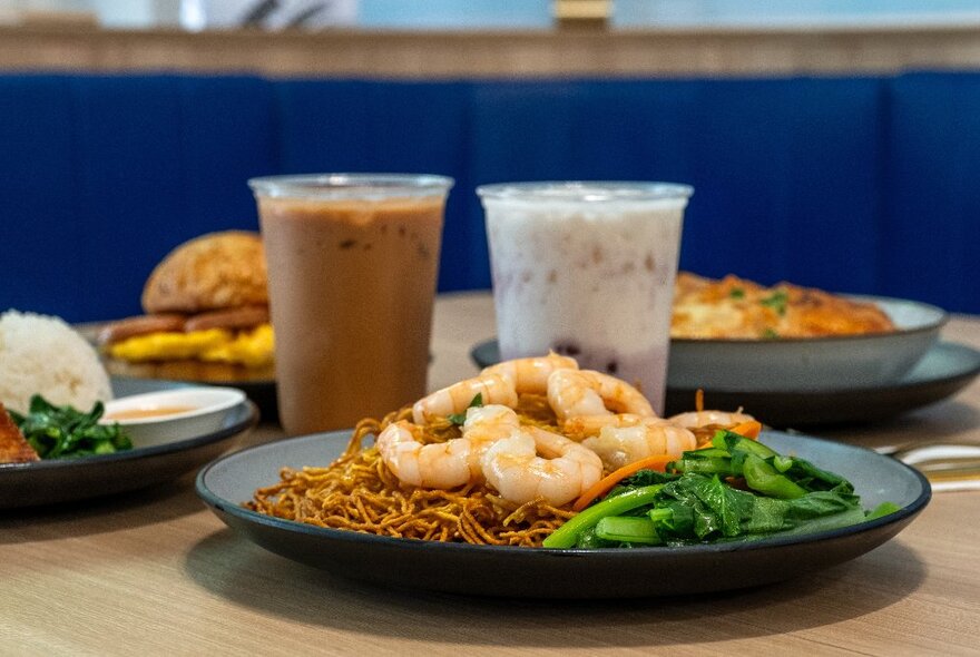 A plate of Hon Kong-style food, with stir-fried noodles, shelled prawns and Chinese broccoli, in front of two iced drinks and other plates of food.