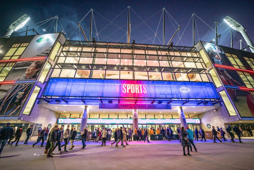 People are gathered outside the MCG sports stadium at night. There is a large neon pink sign on the stadium saying sports.