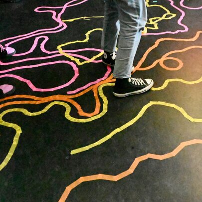 A colourful interactive floor installation or public art project using tape paths on a dark surface, with two sets of legs and feet in the top half of the image.