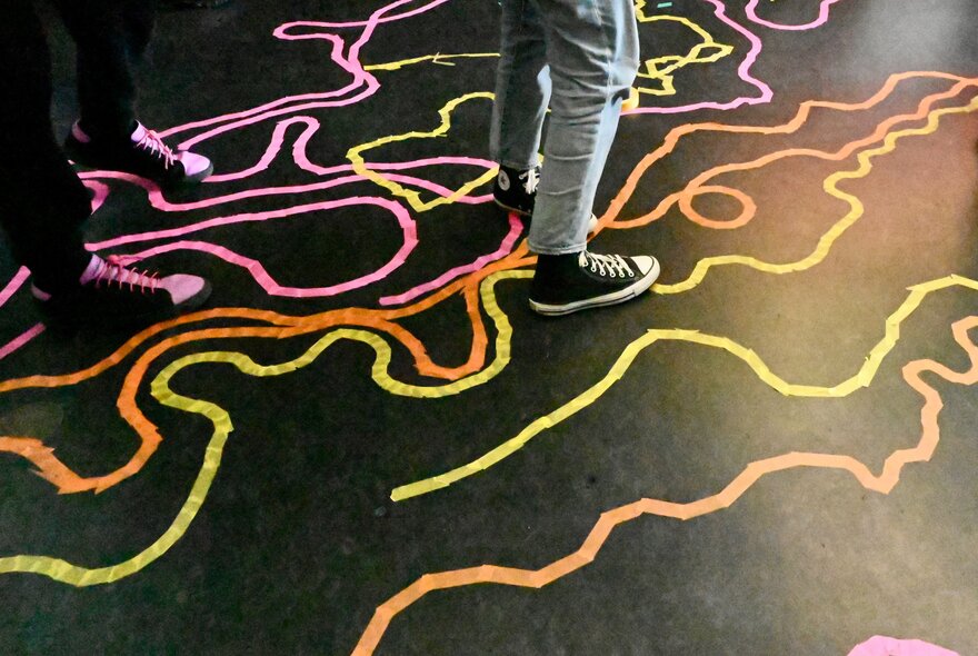 A colourful interactive floor installation or public art project using tape paths on a dark surface, with two sets of legs and feet in the top half of the image.
