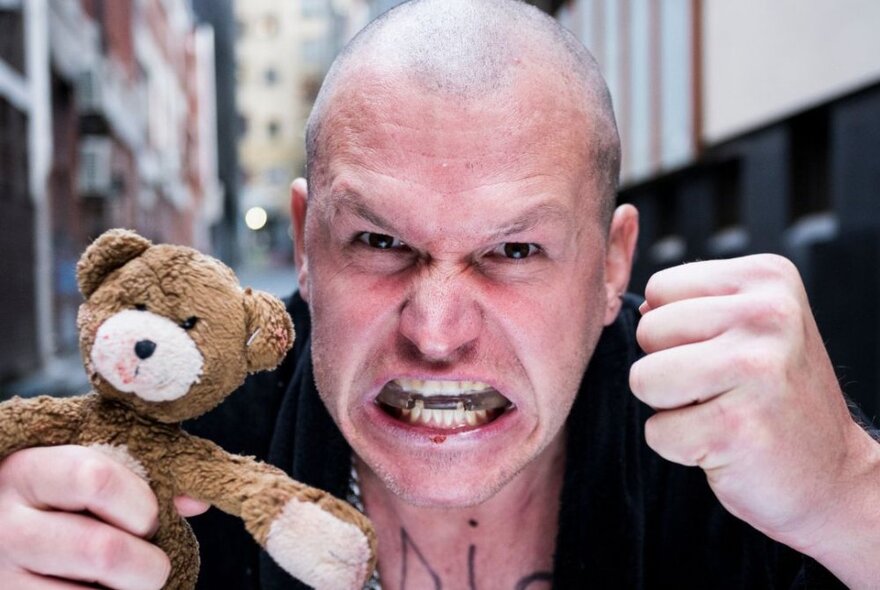 A bald man making an aggressive face, holding a small teddy bear in one of his balled fists, with tattoos on his neck.