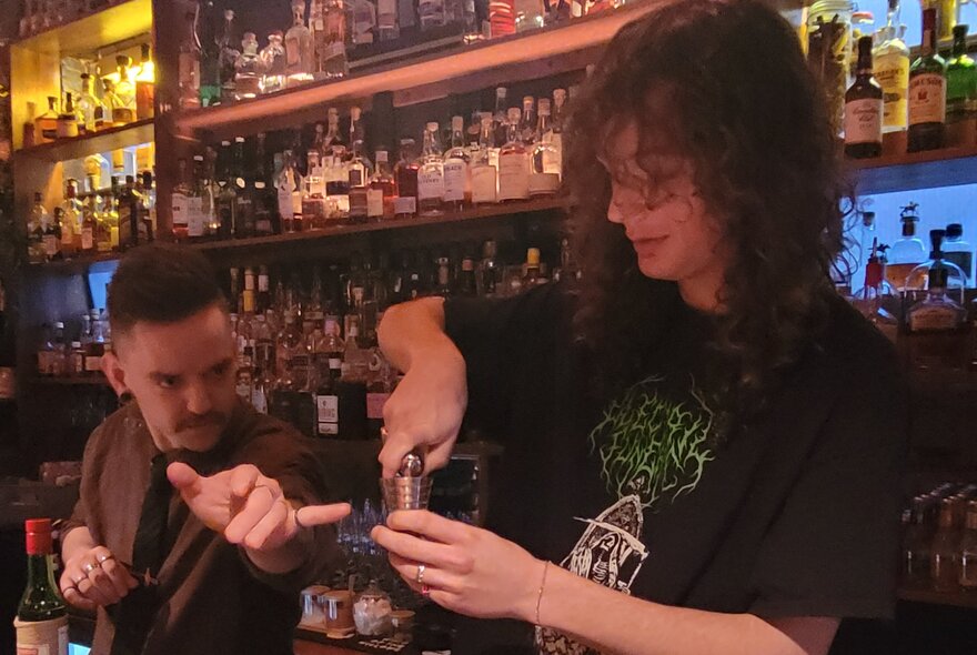 A person making a cocktail behind a bar while being instructed by a bartender.