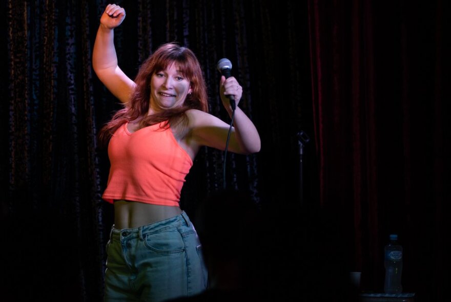 Robyn Reynolds on stage performing her stand-up comedy routine, with both her arms raised in the air and holding a microphone in one hand.