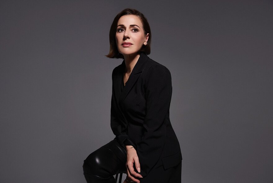 Portrait of renowned musician, Tina Arena, wearing black with sleek, chin length hair and leaning on a stool.