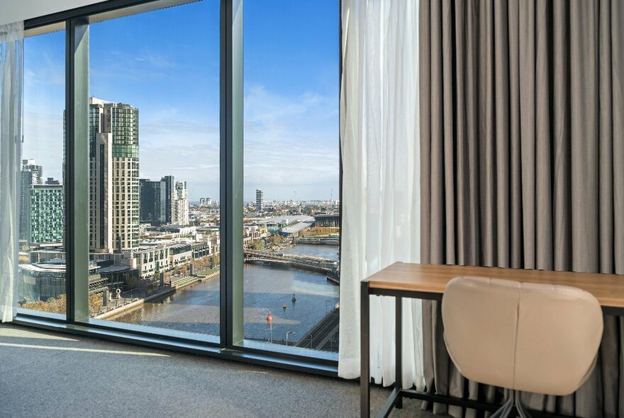 View from tall hotel room window looking onto the Yarra River and Southbank, with beige curtains, table and chair.