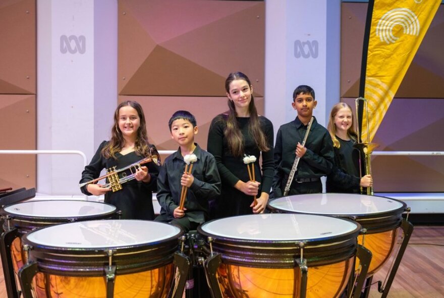 Five young musicians holding instruments or drumsticks, surrounded by four large drums.