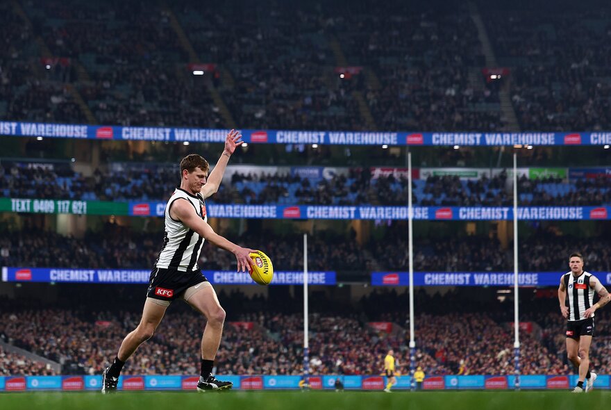 A Collingwood AFL player running in to take a kick holding the yellow football in his right hand; three tiers of fans in the stadium in the background.