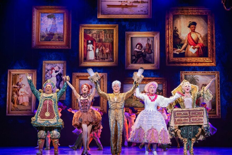 Five members of the cast of Beauty and the Beast in their costumes, on stage holding their hands and raising their arms in the air, the backdrop being a room filled with portraits on a blue wall.