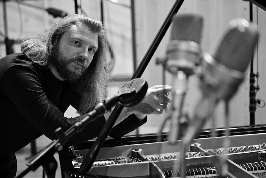 A musician with flowing long hair, leaning against an open grand piano with microphones in the foreground.