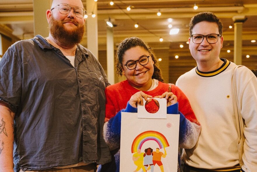 Three people stopping for a picture at an indoor market, the woman in the middle holding up a picture of characters and a rainbow. 