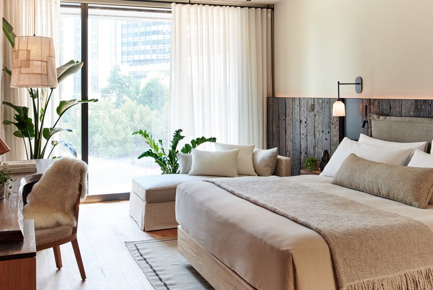 A room at 1 Hotel Melbourne in neutral tones, with large floor to ceiling windows, a king size bed and chaise lounge.