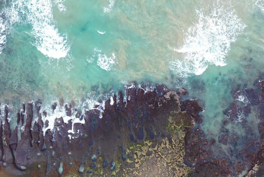 An aerial view of an ocean, showing breaking waves and reefs under the water.