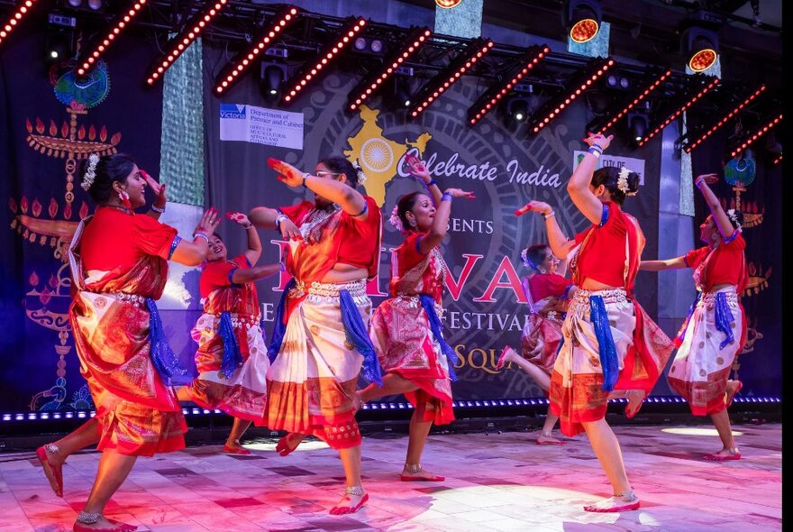 A group of performers dancing on the main stage at Fed Square, all in brightly coloured costumes, celebrating the Festival of Diwali.