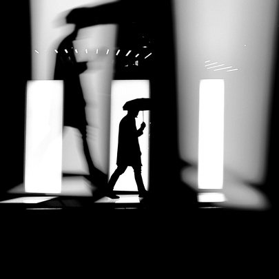 A black-and-white street photograph of a high-contrast scene focused on silhouettes and shadows. 