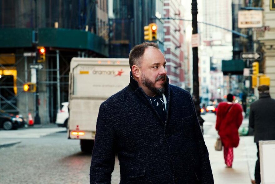 Baritone singer Matthias Goerne, a middle aged man with short hair and a beard, wearing a black overcoat and standing in a busy city street, with buildings, vehicles and pedestrians in the background.