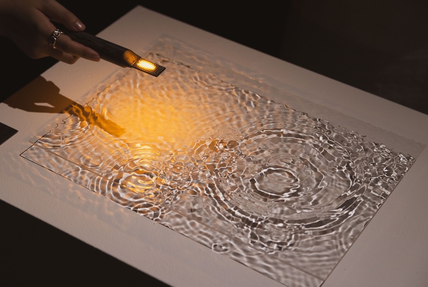A hand holds a small, lit torch over a textured surface with engraved concentric circles and ripples, creating a dramatic lighting effect.