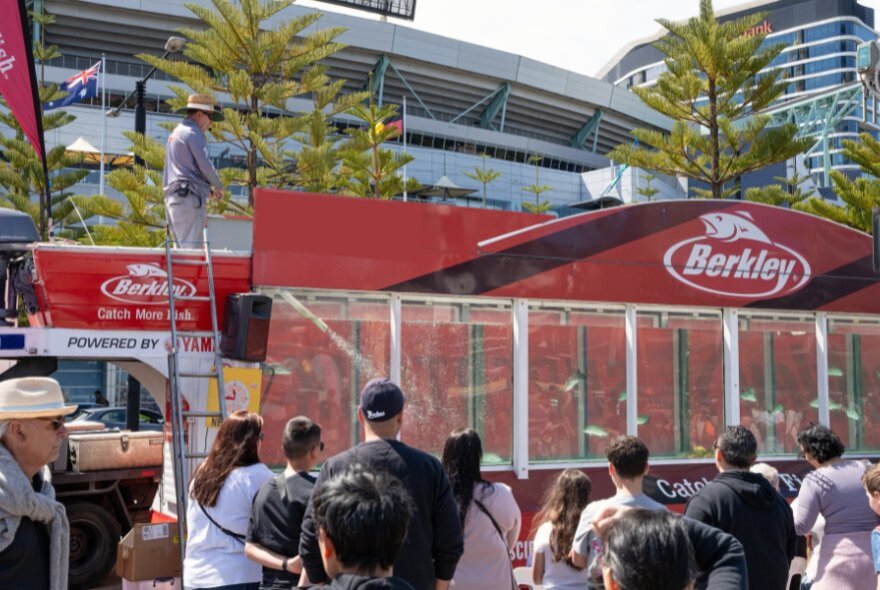 Crowds watching an angler demonstrating fishing tactics at a large tank filled with fish, outdoors at the Melbourne Boat Show.
