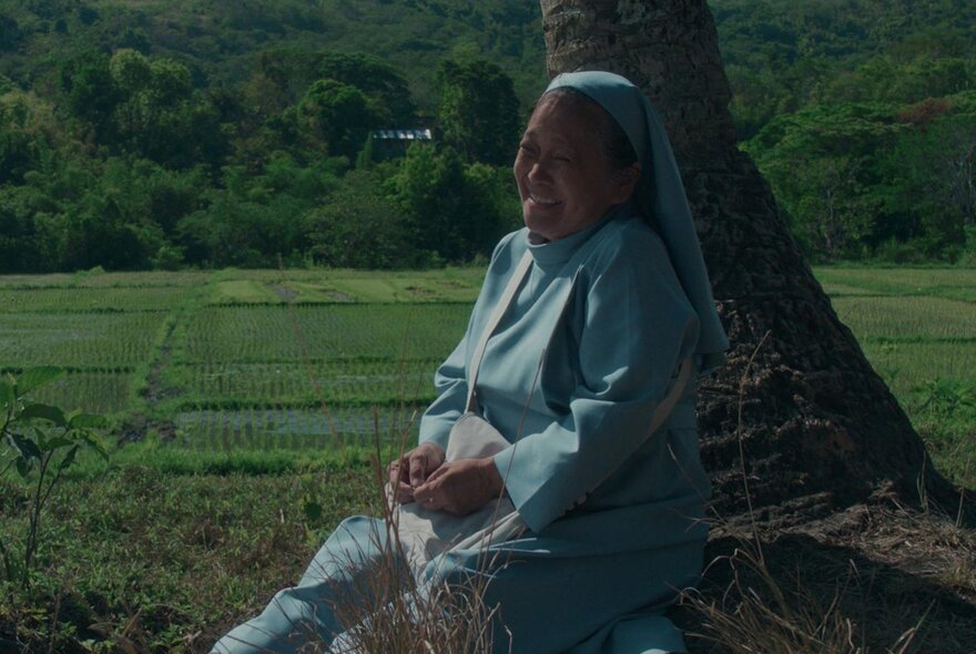 Dark image of a person seated beneath a tree, wearing pale blue, in front of fields.