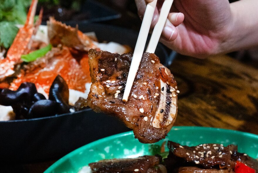 Chopsticks holding up a slice of Korean beef, other dishes visible in the background. 