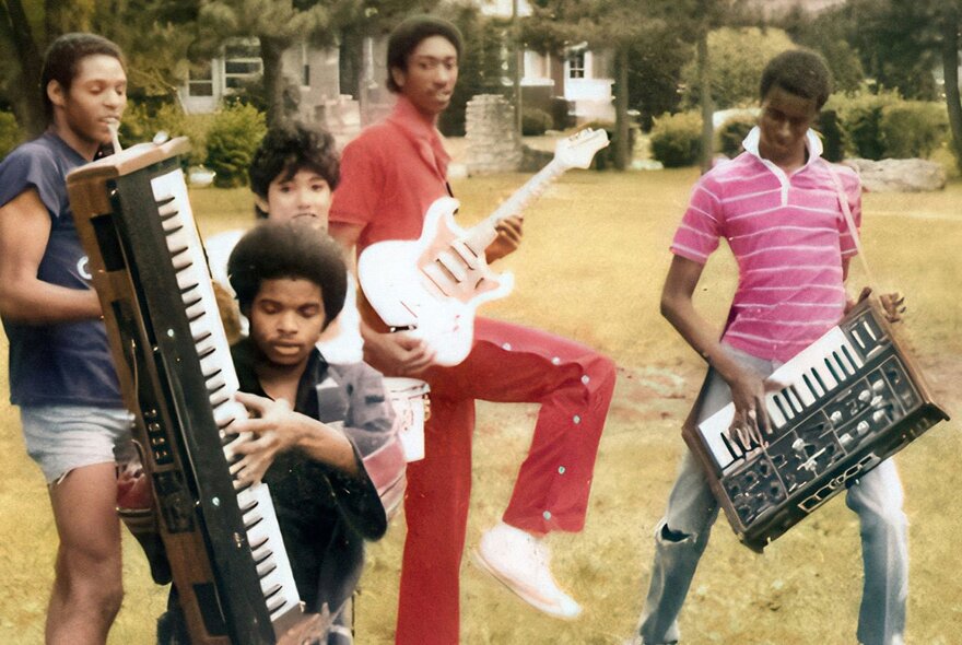 A group of young musicians fooling around with instruments in a park setting, one kid holding a keyboard upways.
