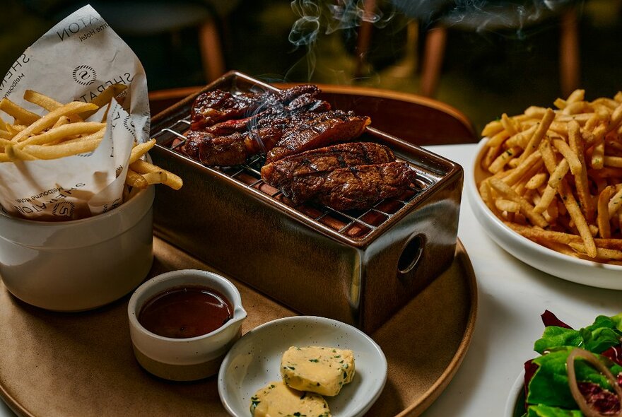 Bowls of chips and condiments next to two pieces of meat steaming on a griddle on a tray.