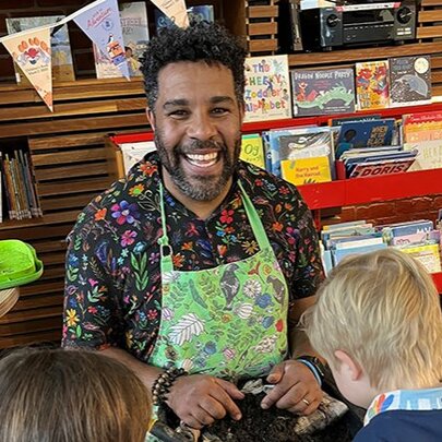 An adult wearing a smock and smiling while interacting with children during a workshop in a library setting.