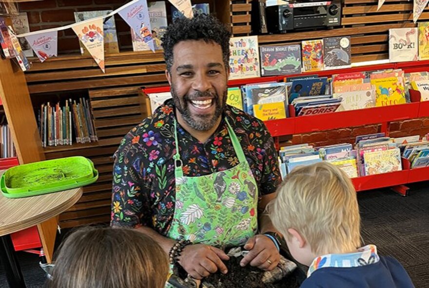 An adult wearing a smock and smiling while interacting with children during a workshop in a library setting.