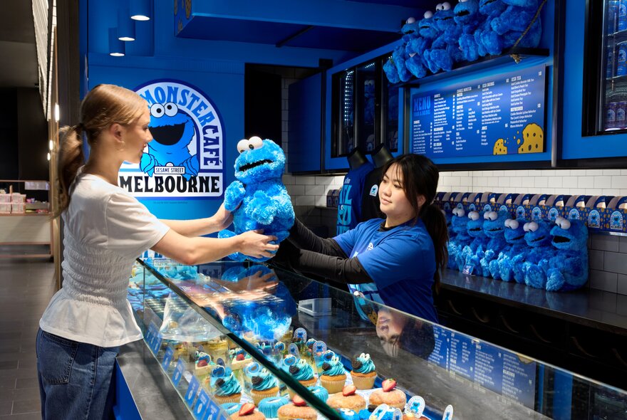A person being handed a large blue Cookie Monster soft toy over the glass counter at the Cookie Monster cafe kiosk at Melbourne Central.