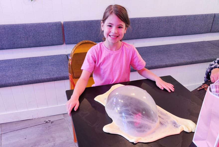 A smiling young girl seated as a table in front of a pile of slime.