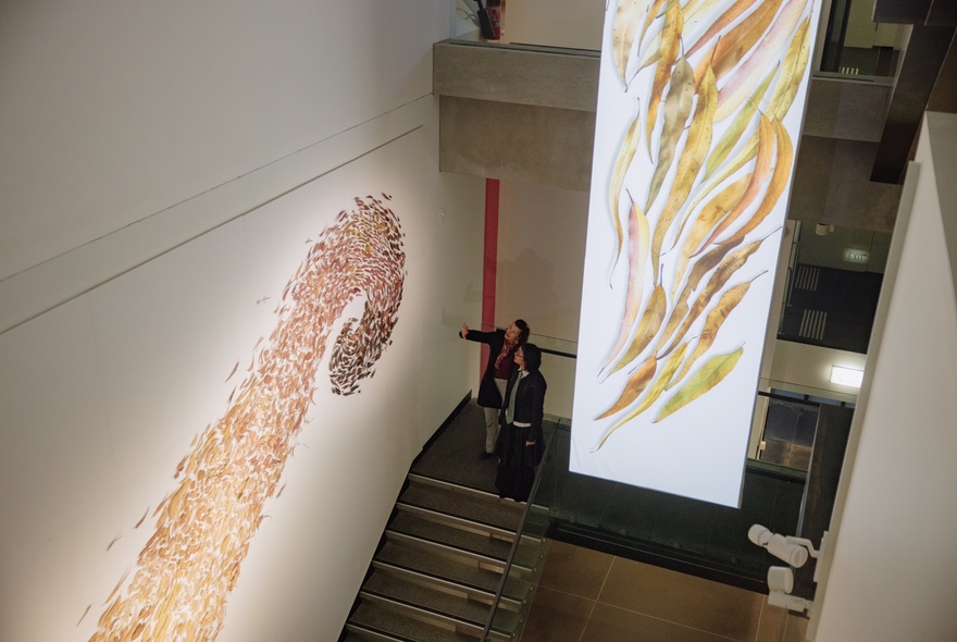People viewing large-scale artworks from a mezzanine staircase in a gallery space.