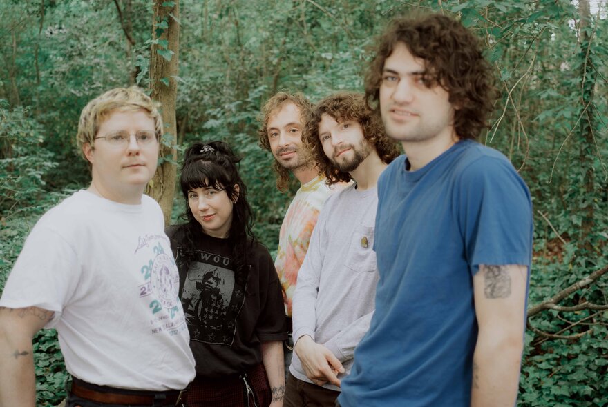 The five members of the indie rock band Wednesday, all looking at the camera, slightly smiling, standing outdoors in a forest setting.