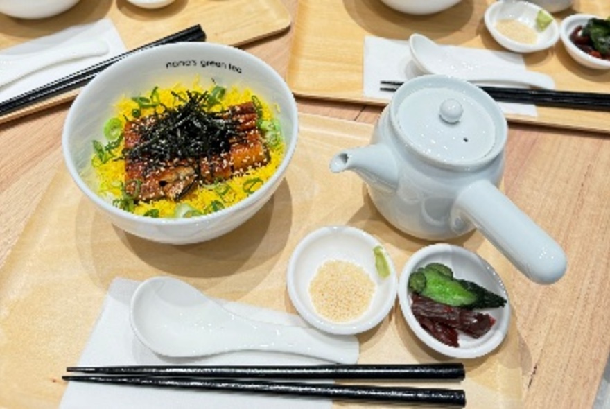 A wooden table with a bowl of food and small side dishes accompanying, a napkin and chopsticks resting in the foreground.