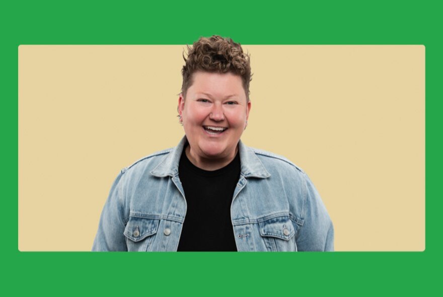 Comedian Kirsty Webeck smiling in a black t-shirt and denim jacket, with short hair and posed against a pale background.