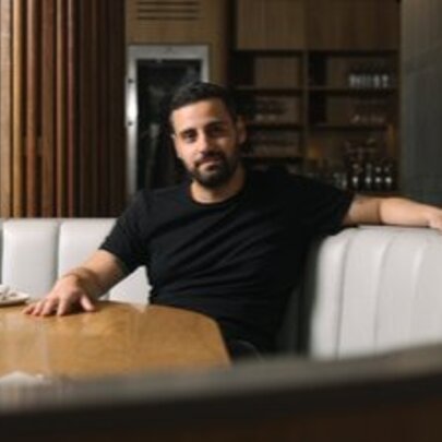 Bearded man seated on a curved beige couch with his hand on a wooden table.