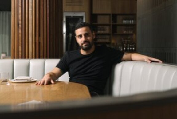 Bearded man seated on a curved beige couch with his hand on a wooden table.