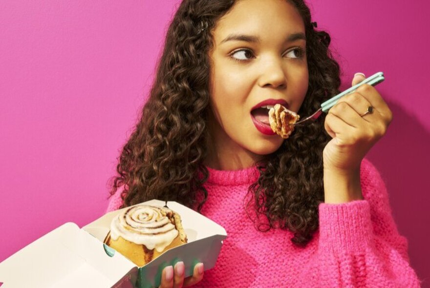 A young woman with darky curly hair wearing a bright pink jumper, about to take a bite of cinnamon bun from a fork she holds close to her mouth, holding a takeaway container with a cinnamon bun in her other hand.