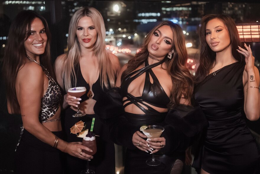 Four women in tight fitting dresses with long hair, posing for a photo while holding drinks.