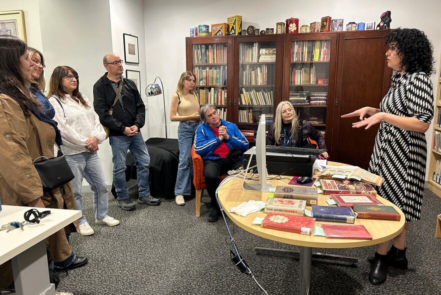 A group of people seated in a carpeted room that is filled with bookcases and books, listening to a tour group host talking.