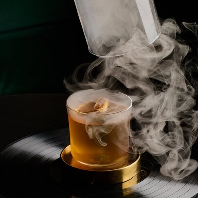 An amber coloured cocktail with a cloche being removed and smoke swirling around it. 