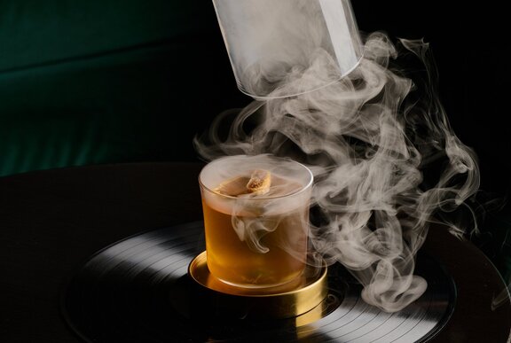 An amber coloured cocktail with a cloche being removed and smoke swirling around it. 