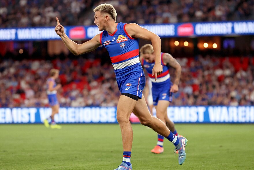 Two Western Bulldogs AFL football players  running on the grass of a football stadium, with spectators watching from the stands.