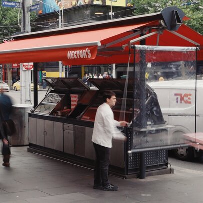 A kiosk in the middle of a city with a red awning and people strolling by. 