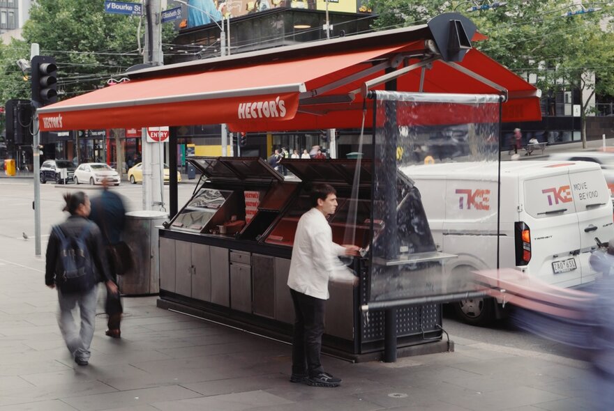 A kiosk in the middle of a city with a red awning and people strolling by. 