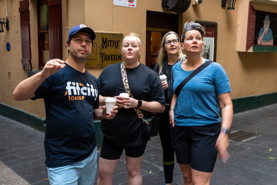 Three people on a walking tour with a Fit City tour guide, each looking up at a building; the peach-coloured Mitre Tavern behind them.