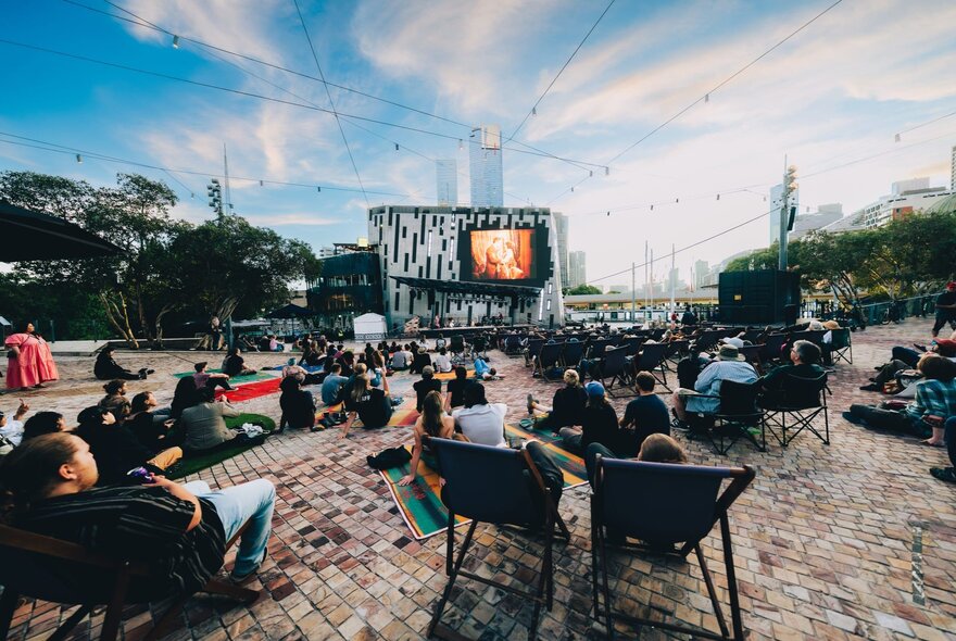 A crowd of people seated on deck chairs, beanbags and picnic rugs, watching an open air film screening at Melbourne's Fed Square.