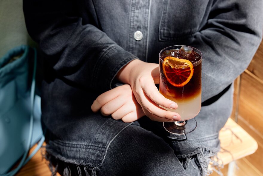 A layered coffee drink in a tall stemmed glass with a slice of orange.