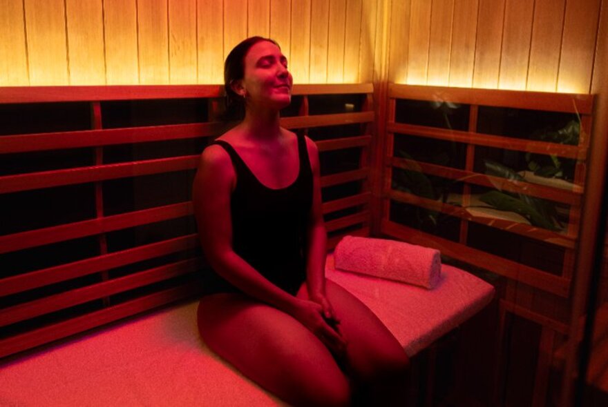 An individual relaxing inside a wooden infrared sauna illuminated by warm red light therapy.