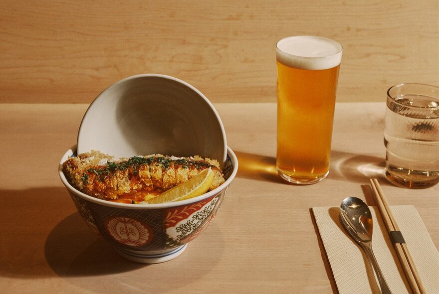 A glass of beer next to a bowl of Japanese food with cutlery.
