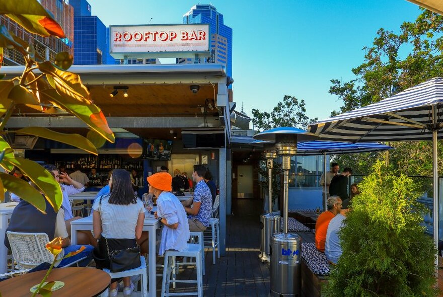 People on a sunny rooftop bar with umbrellas and trees.